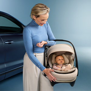Eine Frau in einem blauen Oberteil hält ein Baby in einer beigen Babyschale und steht neben einem dunkelblauen Auto.