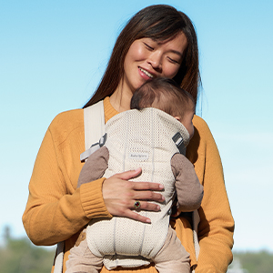 Eine Frau lächelt auf einen Säugling hinab, den sie in einer beigen Babytrage trägt. Sie trägt einen gelben Pullover, und der Himmel ist strahlend blau.