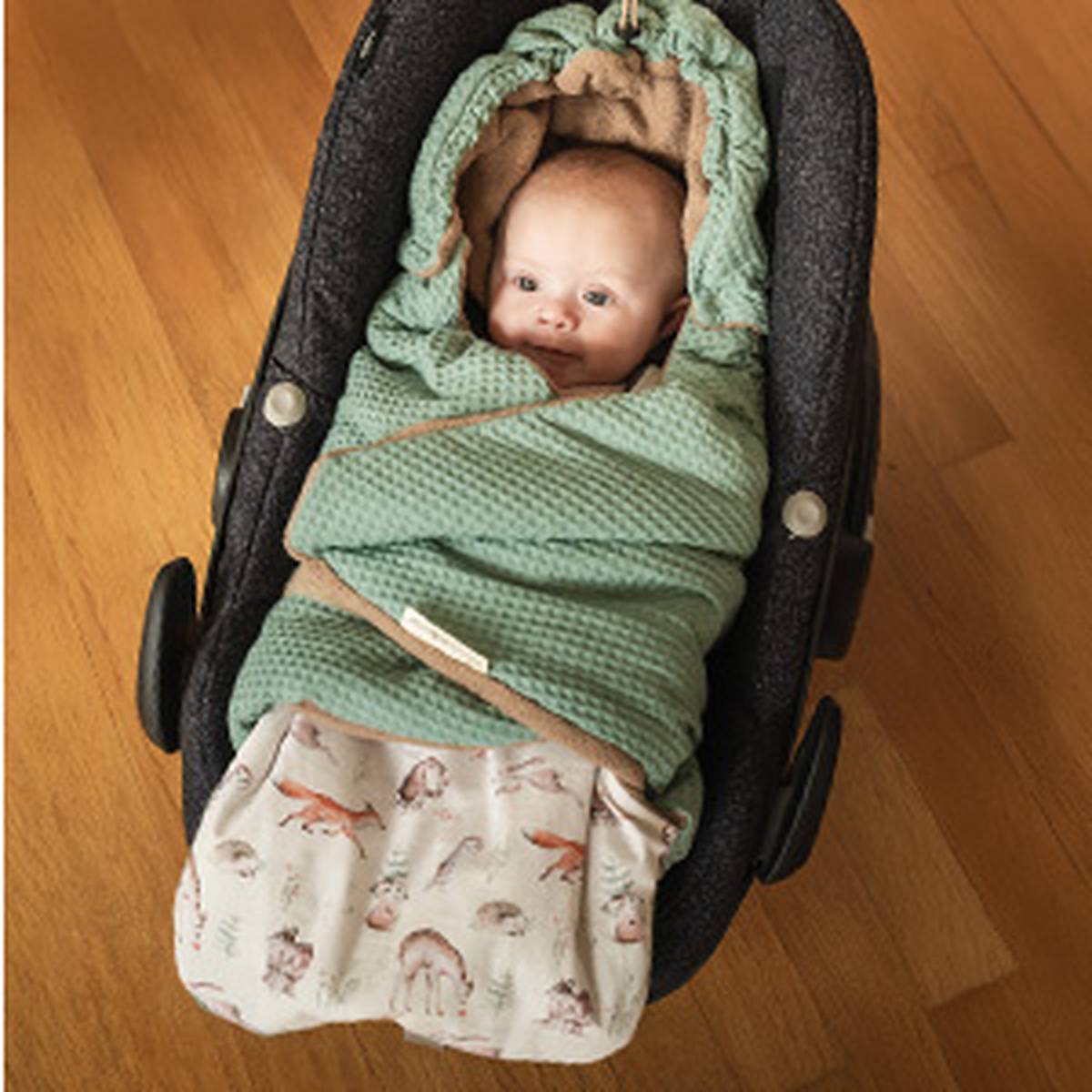 Ein Baby in einem Autositz, eingewickelt in eine grüne Decke mit Waffelmuster und Kapuze, sowie einem Bezug mit Tiermotiven, auf einem Holzboden.