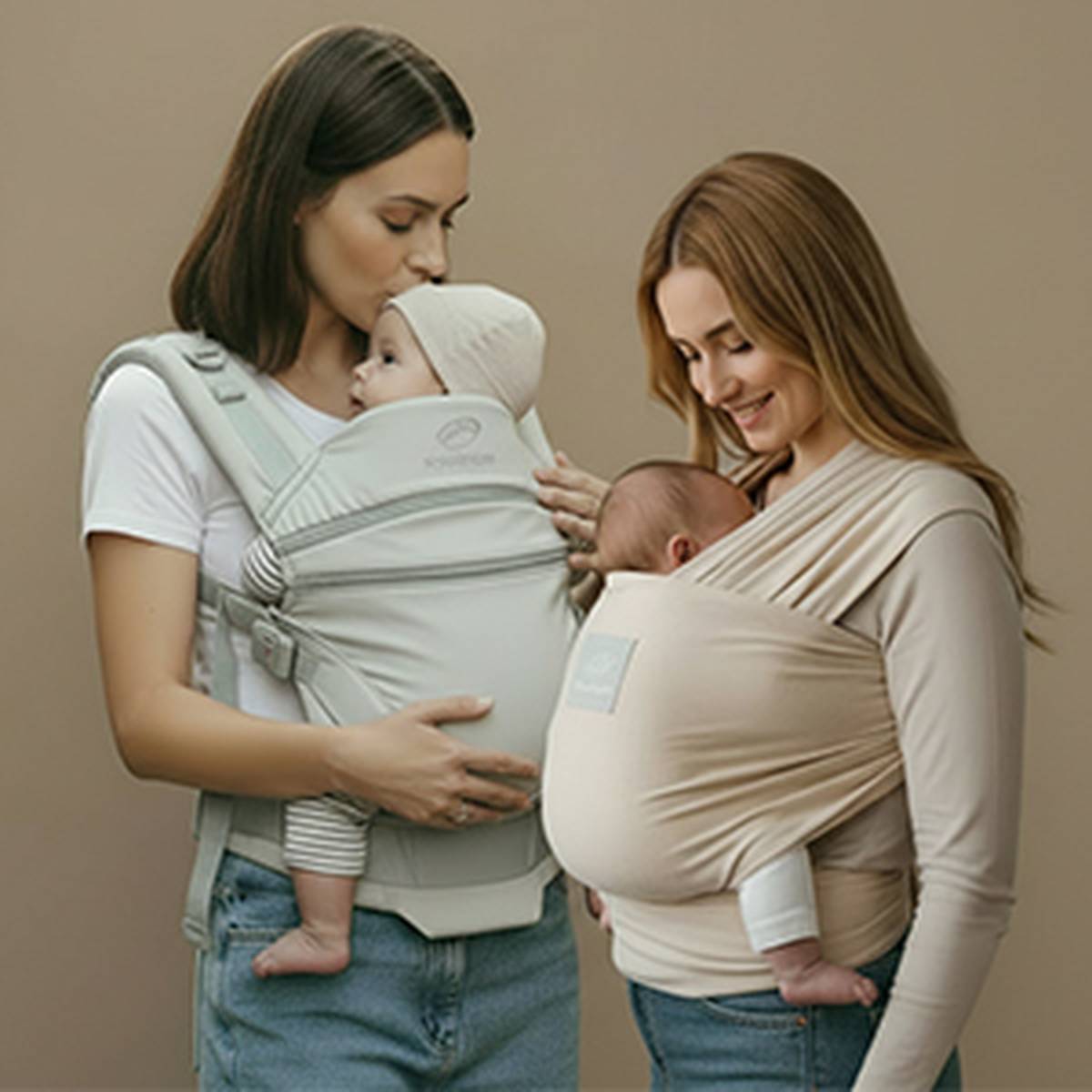 Zwei Frauen, die Säuglinge in Babytragen halten; eine küsst den Kopf eines Babys, beide lächeln und tragen Freizeitkleidung vor neutralem Hintergrund.