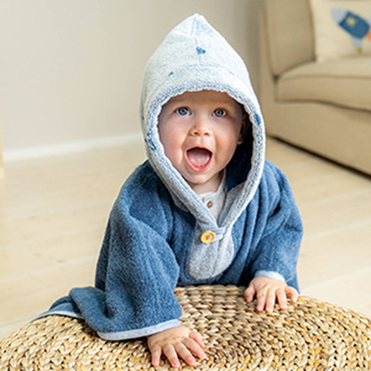 A happy baby wearing a blue hooded bathrobe leans forward on a woven ottoman, with a beige couch in the background.