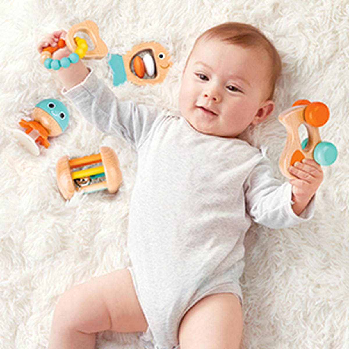 Baby in white onesie lying on a soft surface, holding colorful toys, surrounded by additional toys, smiling.