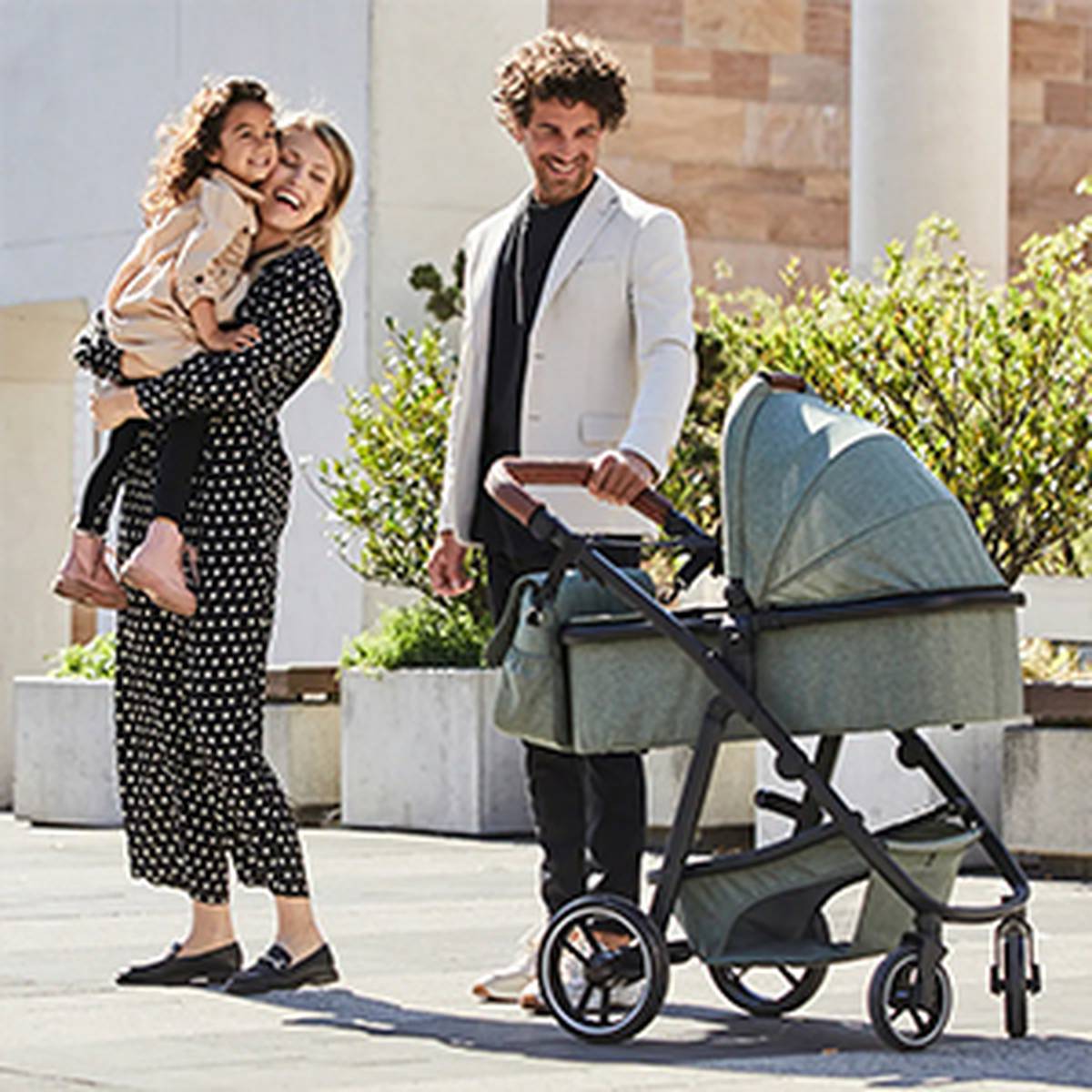 Ein lächelndes Paar mit einem kleinen Kind und einem Kinderwagen steht an einem sonnigen Tag im Freien, umgeben von Grün und moderner Architektur.