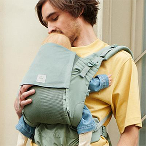 Ein Mann in einem gelben Hemd hält behutsam ein schlafendes Baby in einer grünen Trage und schenkt ihm eine zärtliche, fürsorgliche Umarmung.