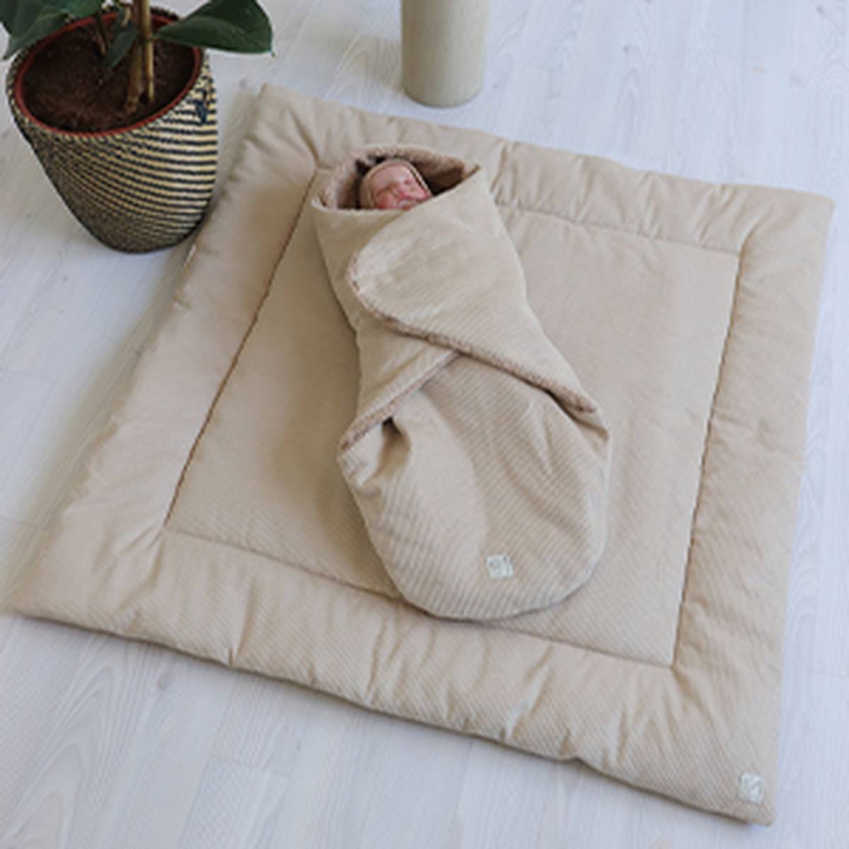 A baby wrapped in a beige swaddle on a matching padded mat, placed on a light wooden floor beside a potted plant.