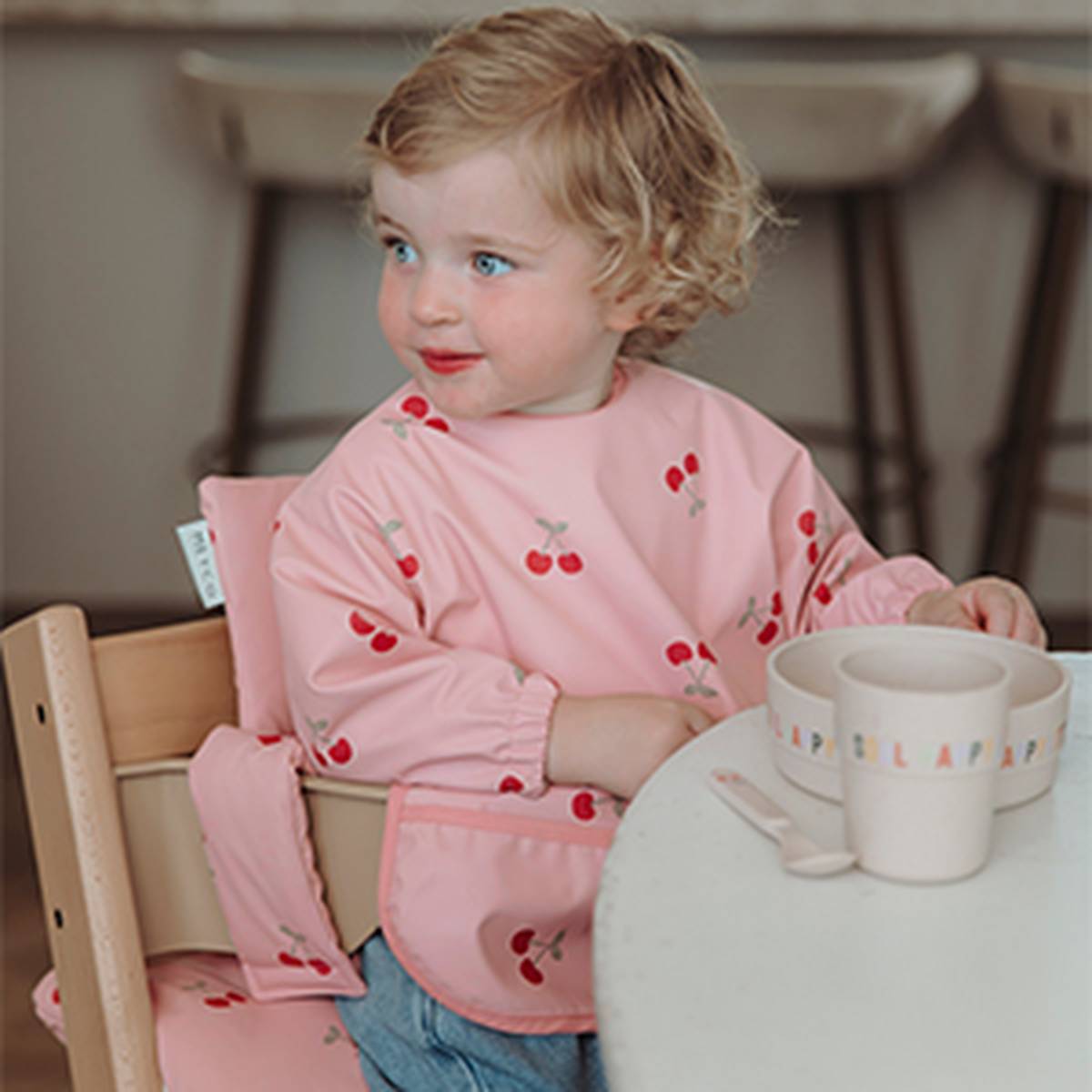 Ein Kleinkind mit blonden Locken sitzt in einem Hochstuhl und trägt ein rosa Lätzchen mit Kirschmuster; neben ihm stehen eine Schüssel und ein Becher.