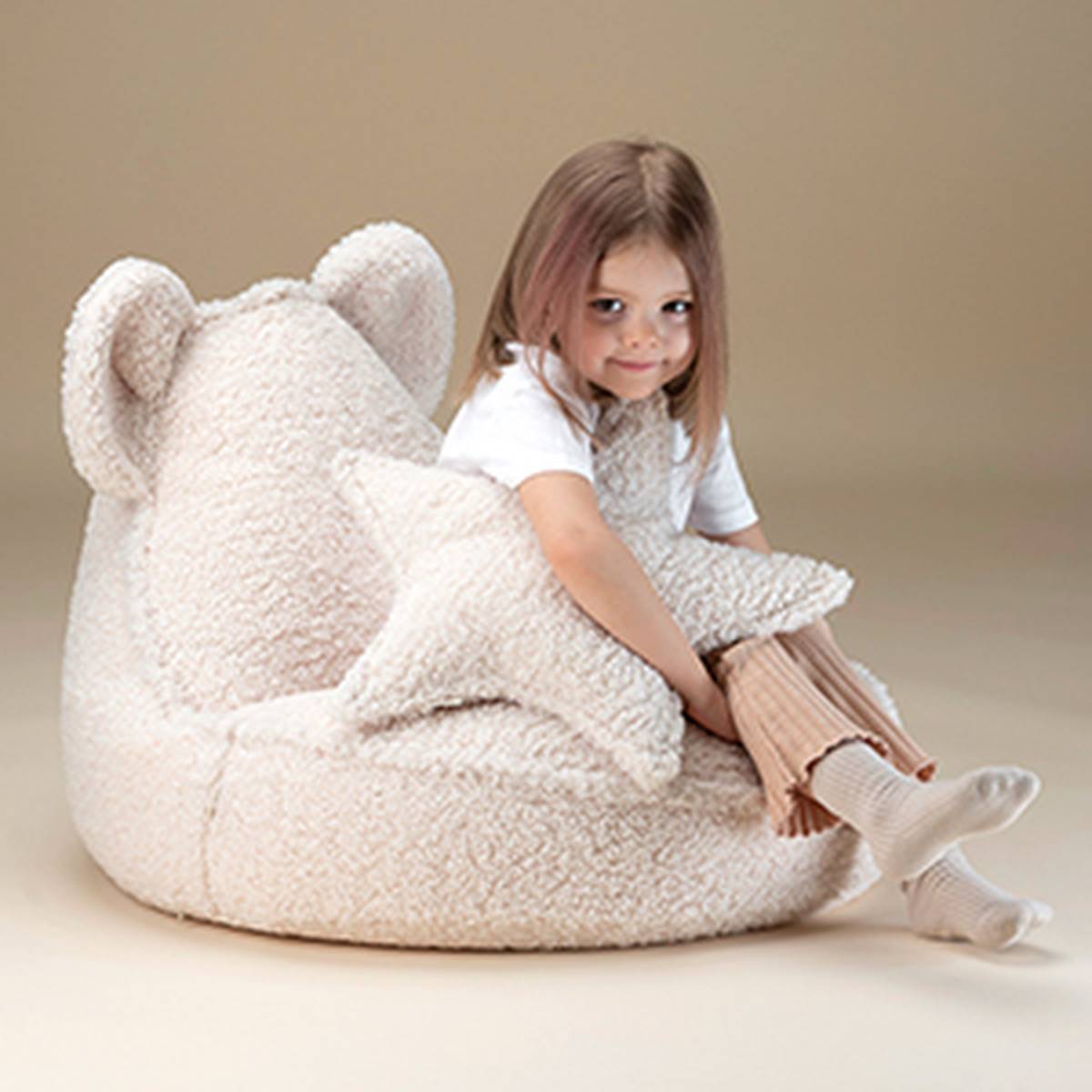 A young girl sitting on a fluffy, bear-shaped bean bag chair, hugging a matching plush star-shaped pillow in a neutral toned room.