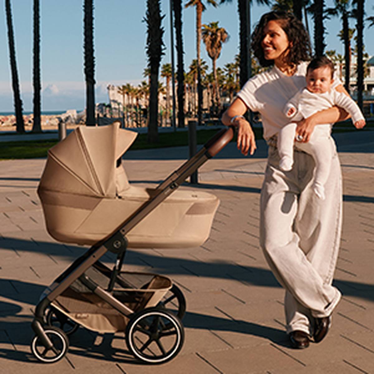 Eine Frau steht lächelnd mit einem Baby im Arm neben einem beigefarbenen Kinderwagen auf einer sonnigen Promenade, im Hintergrund sind Palmen zu sehen.