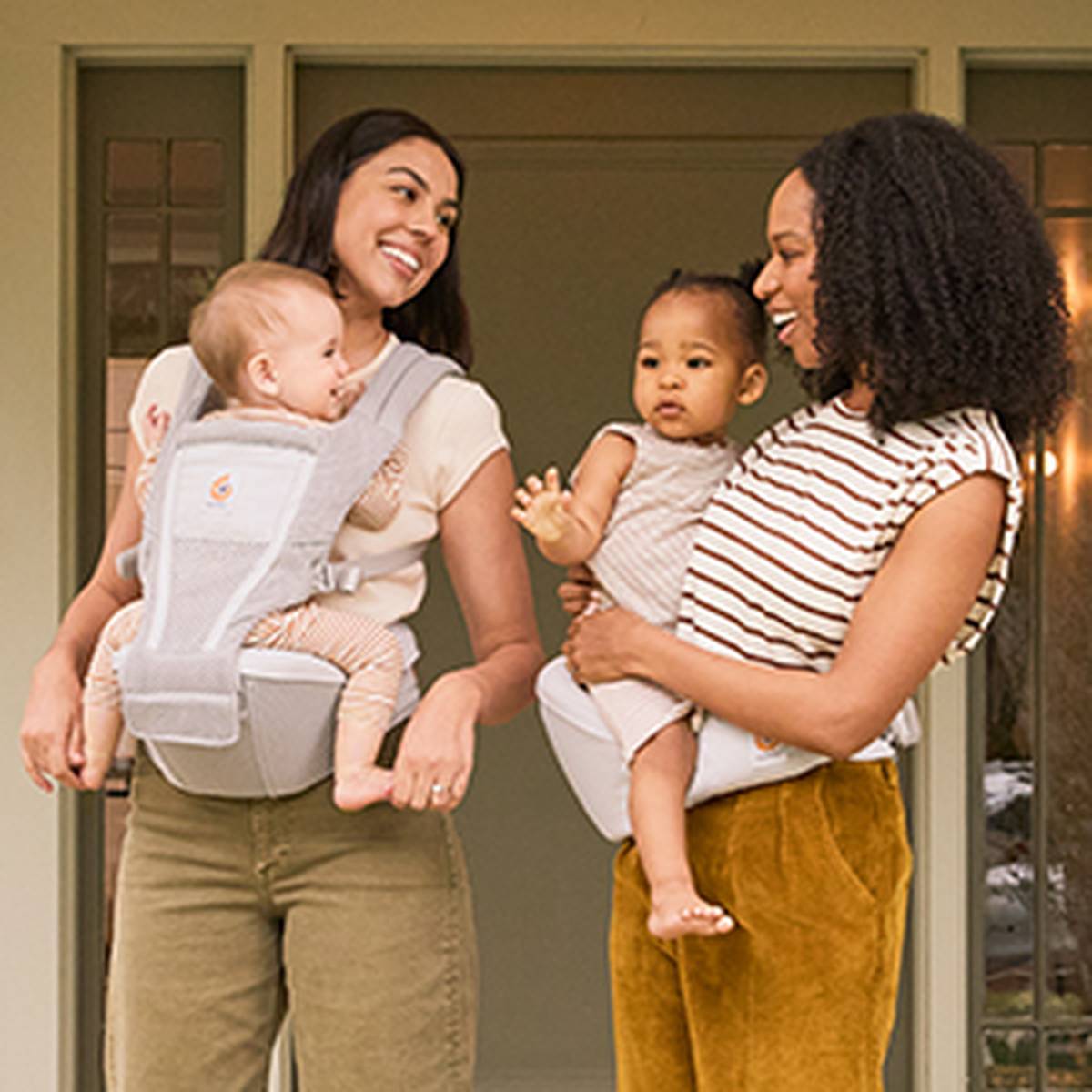 Zwei Frauen, die sich anlächeln und jeweils ein Baby in einer weichen Trage halten, stehen vor einem Haus.