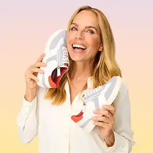 Lächelnde Frau in einer weißen Bluse hält ein Paar Turnschuhe vor ihrem Gesicht vor einem pastellfarbenen Hintergrund.