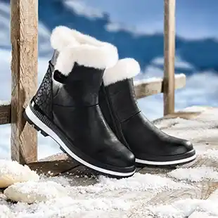Schwarze Winterstiefel mit weißem Fellfutter auf einer verschneiten Holzoberfläche, im Hintergrund eine verschneite Landschaft.