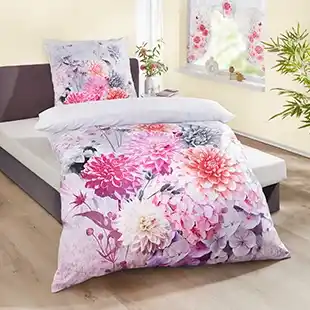 Bett mit Bettwäsche mit Blumenmuster in Rosa- und Violetttönen, neben einem Fenster mit Blumenvorhängen und einer Topfpflanze in der Nähe.