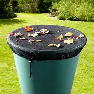 Ein grüner Regenfass mit einer schwarzen Netzabdeckung, übersät mit Herbstblättern, steht auf einer Rasenfläche mit Bäumen im Hintergrund.