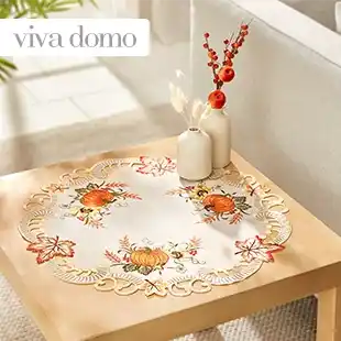 Ein Holztisch mit einer bestickten Tischdecke mit Herbstmotiven wie Kürbissen und Blättern, geschmückt mit zwei weißen Vasen mit Trockenblumen.