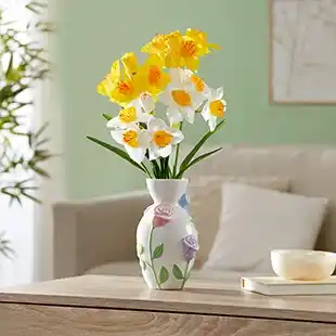 Leuchtend gelbe und weiße Osterglocken in einer dekorativen Vase auf einem Holztisch, aufgestellt in einem gemütlichen Wohnzimmer mit grünen Wänden und einer Pflanze.