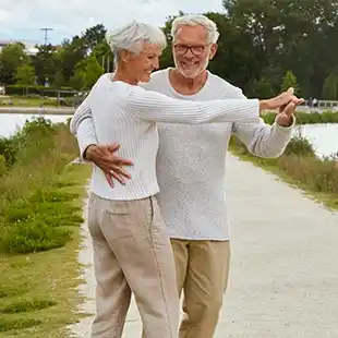 Älteres Ehepaar, das an einem sonnigen Tag in einem Park an einem Fluss lächelt und tanzt, beide tragen helle Pullover und Hosen.
