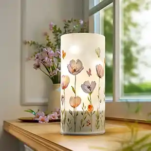 Eine dekorative Lampe mit Blumenmuster auf einem Holztisch am Fenster, umgeben von rosa Blumen und Grünpflanzen in sanftem, natürlichem Licht.