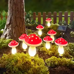 Leuchtende, pilzförmige Gartenlampen mit roten Kappen stehen zwischen Moos und Mulch in der Nähe eines Baumes, im Hintergrund ist ein kleines Solarpanel zu sehen.