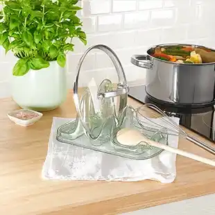 Ein Glasdeckel und ein Löffel liegen auf einer Löffel- und Deckelablage, neben einer Basilikumpflanze, während auf dem Herd ein Topf kocht.