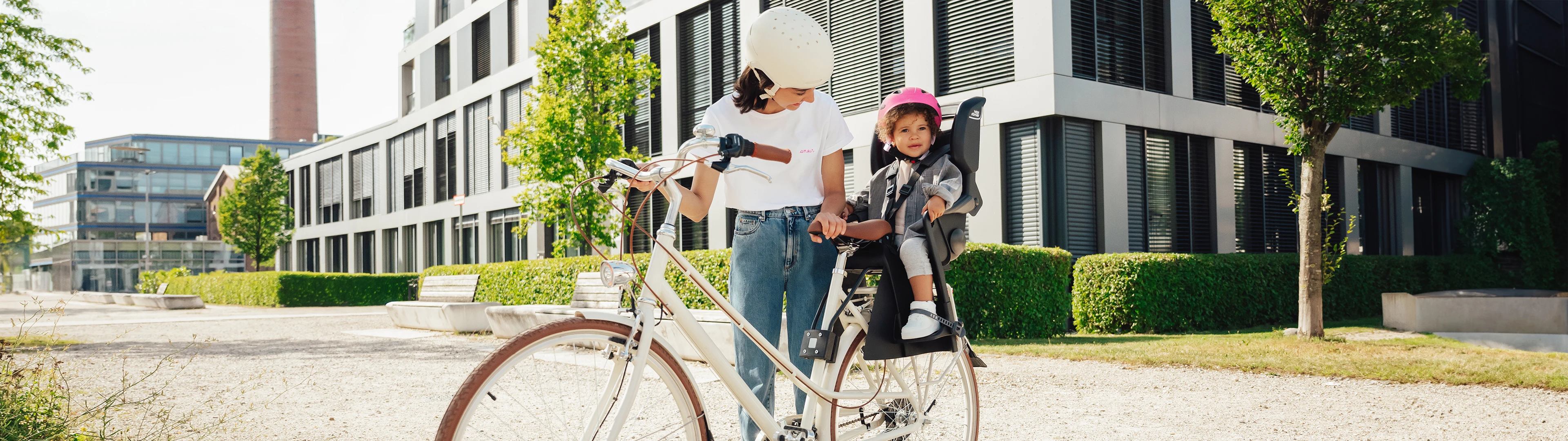 Frau und Kind auf einem Fahrrad in der Stadt, beide mit Helm. Das Kind sitzt hinten auf dem Fahrrad. Im Hintergrund moderne Gebäude und Bäume.
