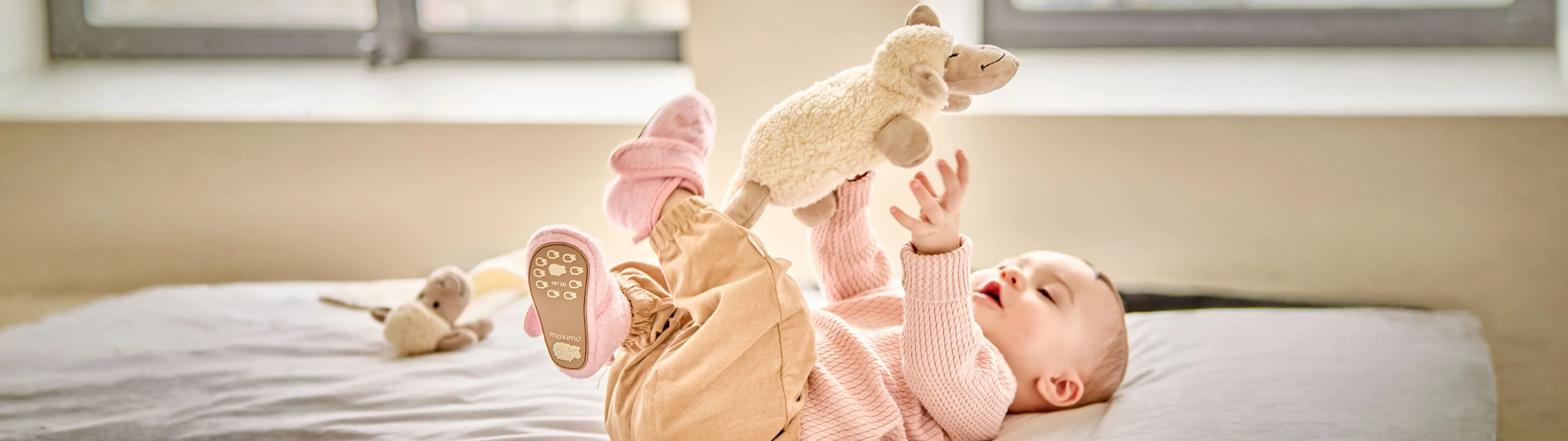 Baby spielt auf einem Bett mit einem weichen Kuscheltier in Schafform und trägt rosa Bekleidung sowie passende Hausschuhe.