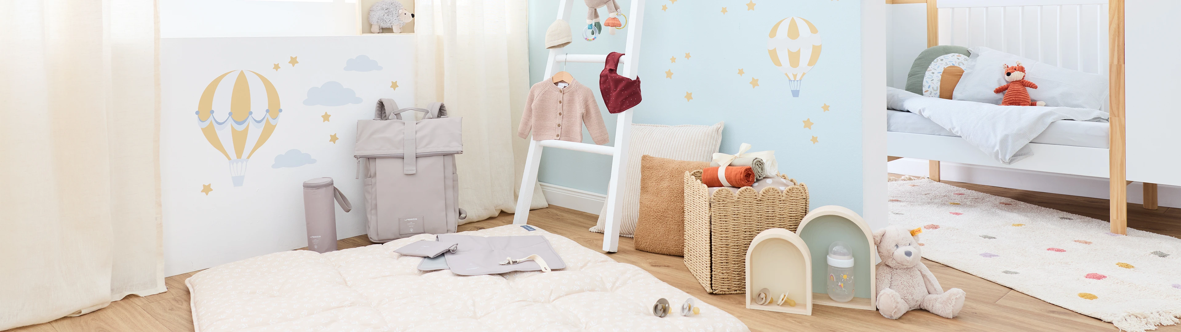 Eine gemütliche Kinderzimmerszene mit einem flauschigen Teppich, Babykleidung auf einer Leiter, einem Rucksack, Fläschchen und Kuscheltieren vor einer pastellfarbenen Wand mit Ballonmotiven.