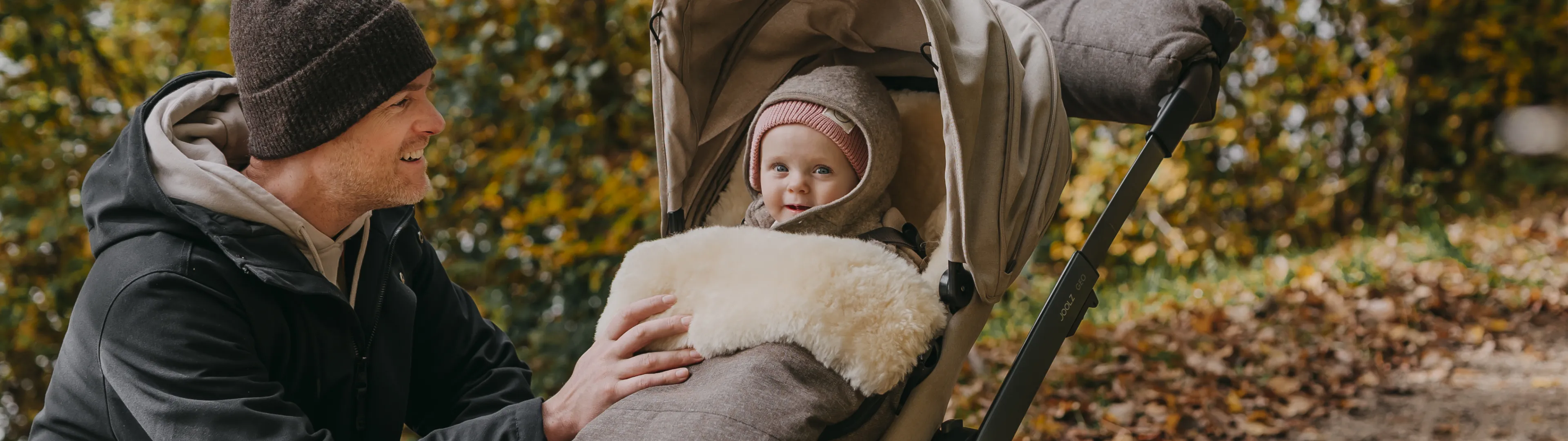 Kinderwagen mit Baby wird auf einem herbstlichen Waldweg genutzt. Das Kind sitzt warm eingepackt im Kinderwagen mit Fußsack und Verdeck, während eine Begleitperson daneben aus der Hocke in den Kinderwagen schaut.