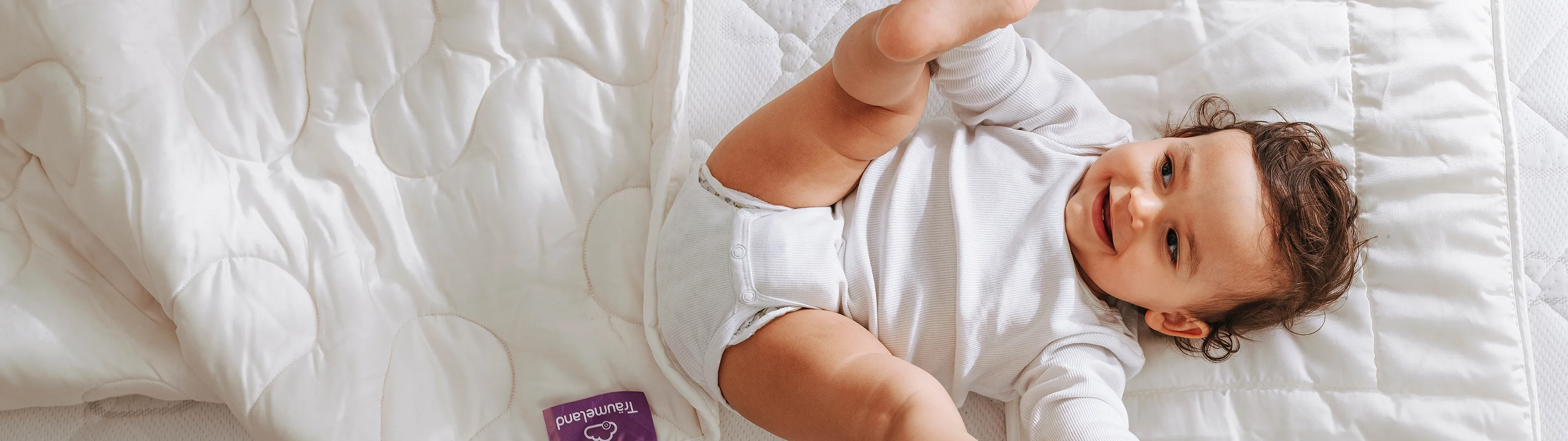 Ein Baby in einem weißen Strampler liegt auf einer gesteppten Matratze, hält spielerisch seine Füße und lächelt; neben ihm liegt eine weiße Decke.