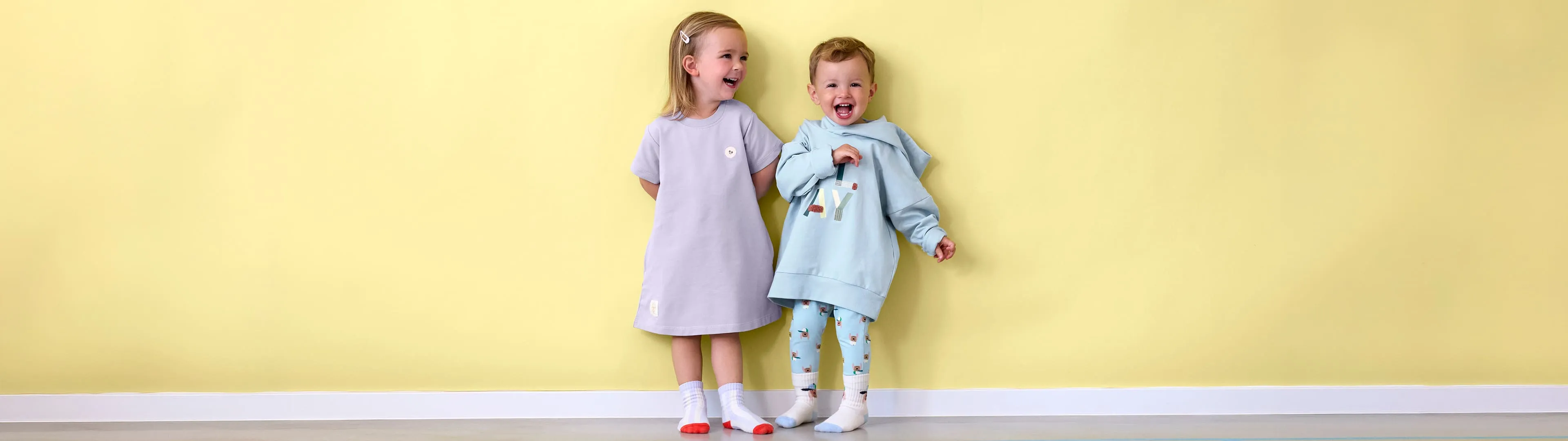 Zwei lächelnde Kinder stehen vor einer gelben Wand; eines trägt ein lilafarbenes Kleid, das andere einen hellblauen Kapuzenpullover sowie eine Leggings – beide tragen Socken.