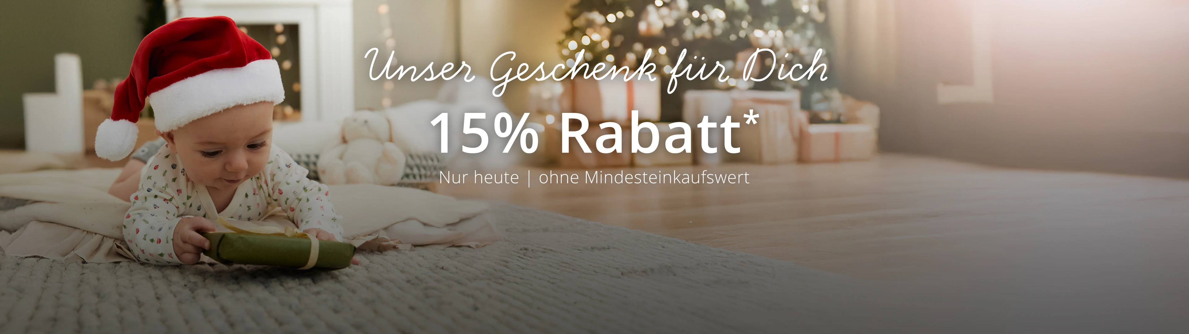 Ein Säugling mit Weihnachtsmannmütze liegt auf dem Bauch und greift nach einem kleinen, grün verpackten Geschenk auf einem strukturierten Teppich; im deutschen Text wird ein Rabatt* von 15 % ohne Mindesteinkaufswert angekündigt.