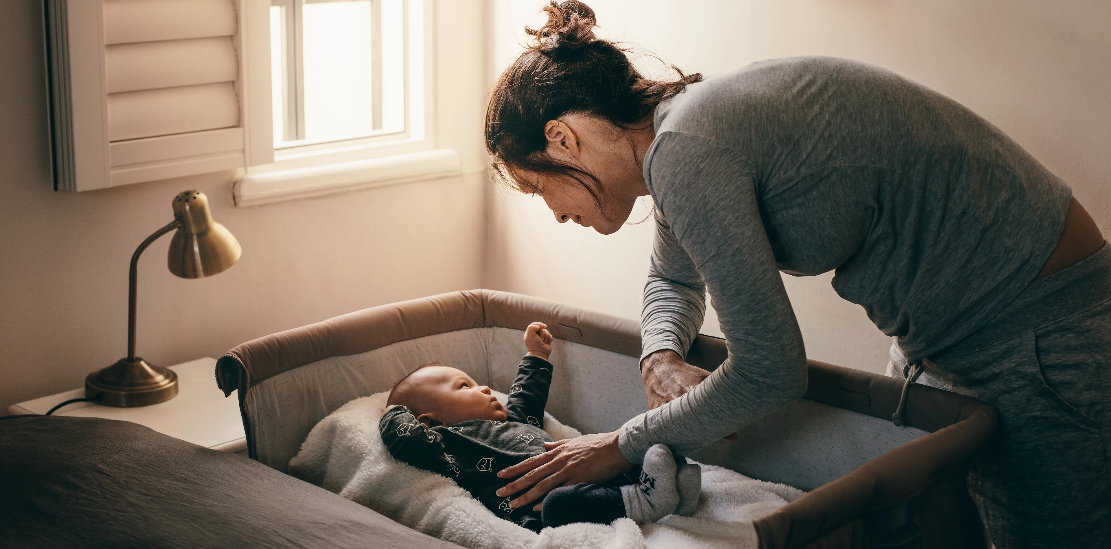 Eine Frau in grauer Kleidung beugt sich über ein Baby in einem Kinderbett und richtet die Decke. Sanftes Licht fällt durch ein Fenster in den Raum.
