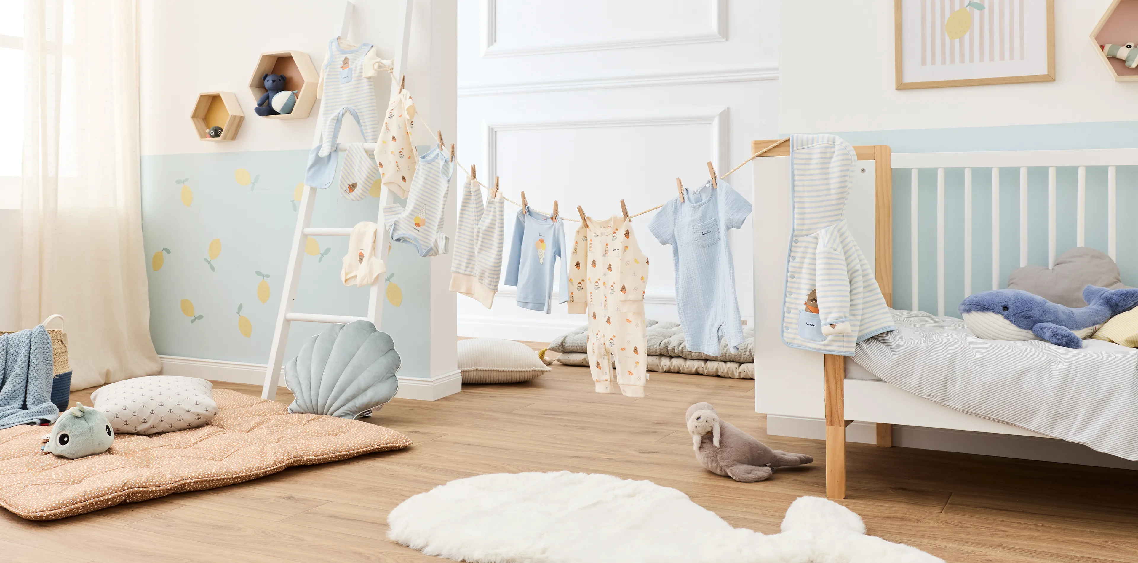 Auf einer weißen Leiter hängen Babykleidungsstücke in Pastellfarben mit Mustern, daneben steht ein Stoffbär. Kissen und sanfte Farben schaffen eine gemütliche Atmosphäre im Kinderzimmer.