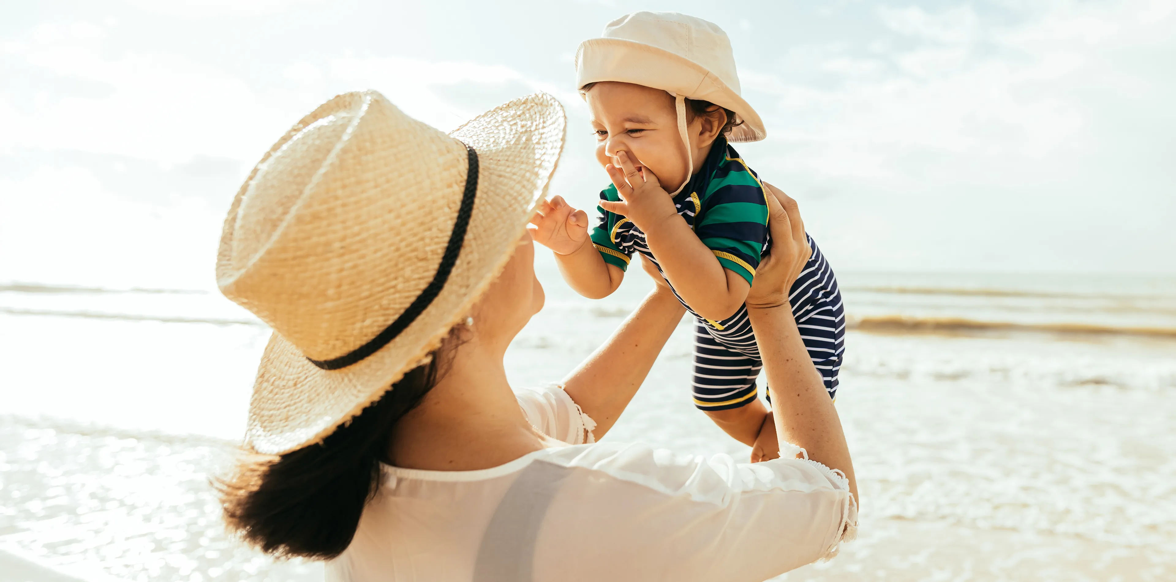 Eine Person mit Strohhut hebt am Strand ein lächelndes Baby mit Sonnenhut und gestreiftem Outfit hoch, im Hintergrund das Meer.