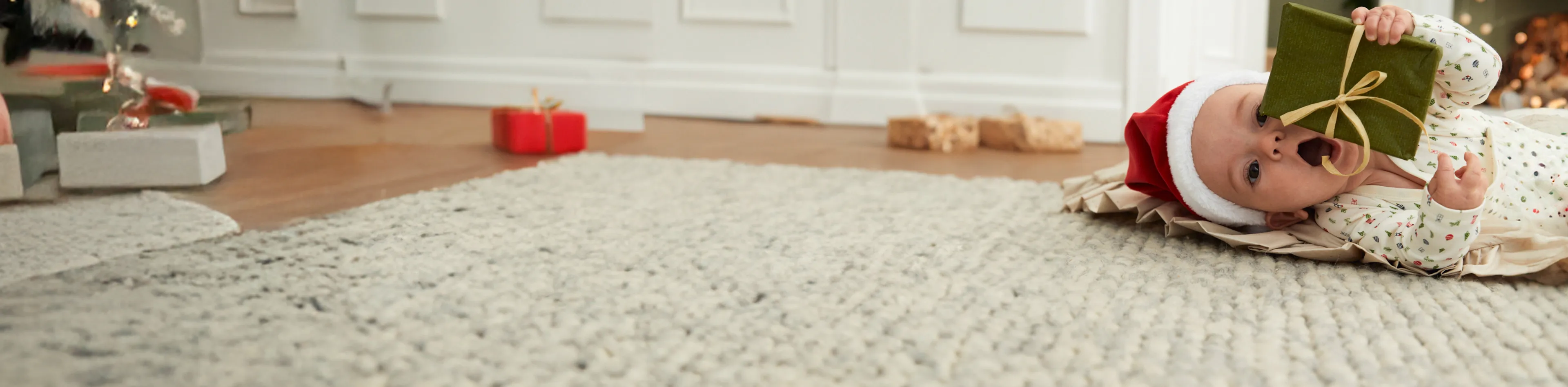 Baby mit Weihnachtsmannmütze liegt auf dem Teppich und hält verspielt ein grünes Geschenk. Im Hintergrund sind ein Weihnachtsbaum und Geschenke zu sehen.