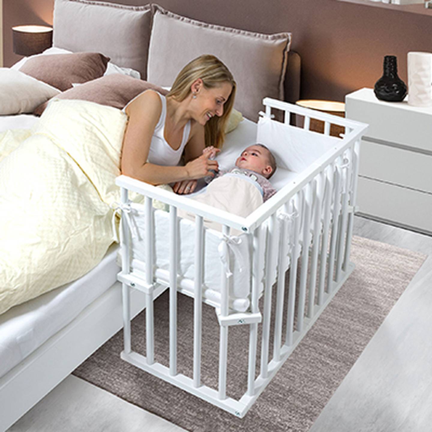 Eine Mutter lächelt ihr Baby an, das in einem weißen Beistellbettchen am Bett befestigt ist und so eine gemütliche und sichere Schlafgelegenheit bietet.