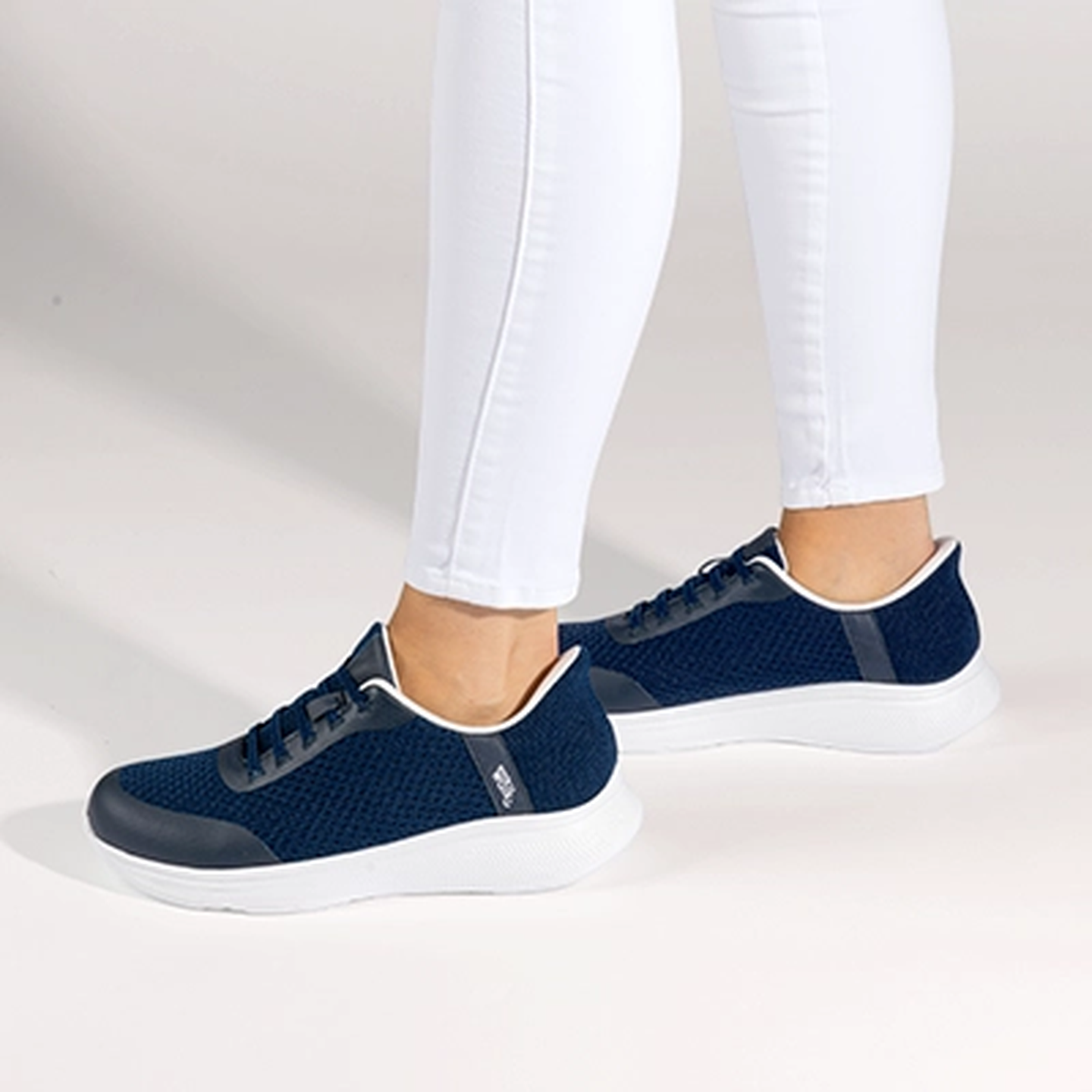 Eine Person in weißen Leggings schlüpft vor einem hellen Hintergrund in ein Paar marineblaue Sneakers mit weißen Sohlen.