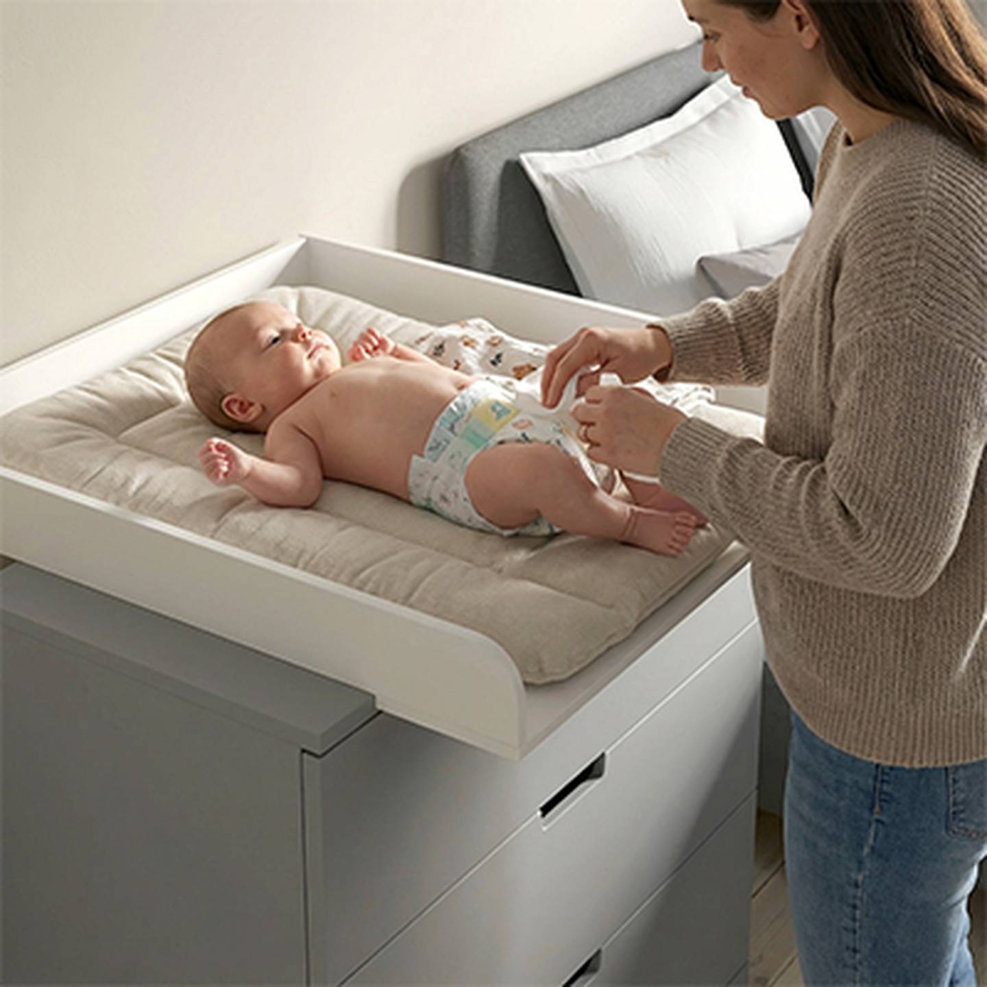 Eine Frau wickelt ein Baby auf einem gepolsterten Wickeltisch in einem Zimmer mit grauen Möbeln und einem Bett im Hintergrund.