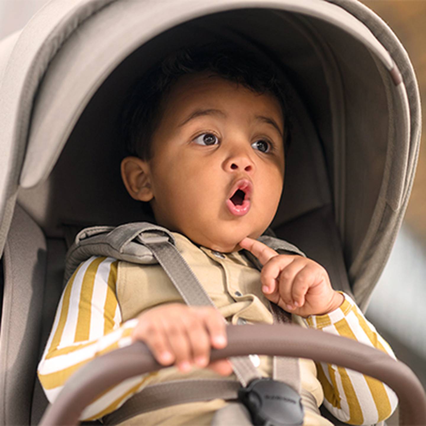 Ein kleines Kind im Kinderwagen blickt überrascht drein – mit weit aufgerissenen Augen und einer Hand nahe am Mund –, bekleidet mit einem gestreiften Hemd und einer beigen Weste.