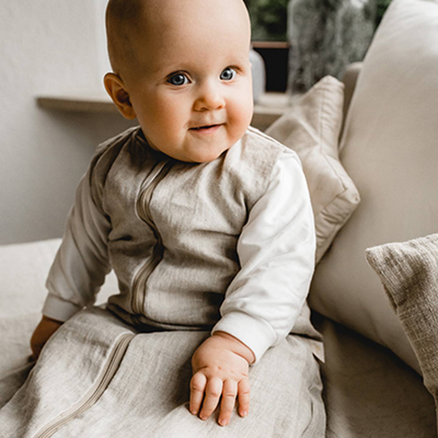 Ein Baby in beiger Kleidung sitzt auf einem Sofa, blickt nach oben und lächelt, umgeben von weißen Kissen.