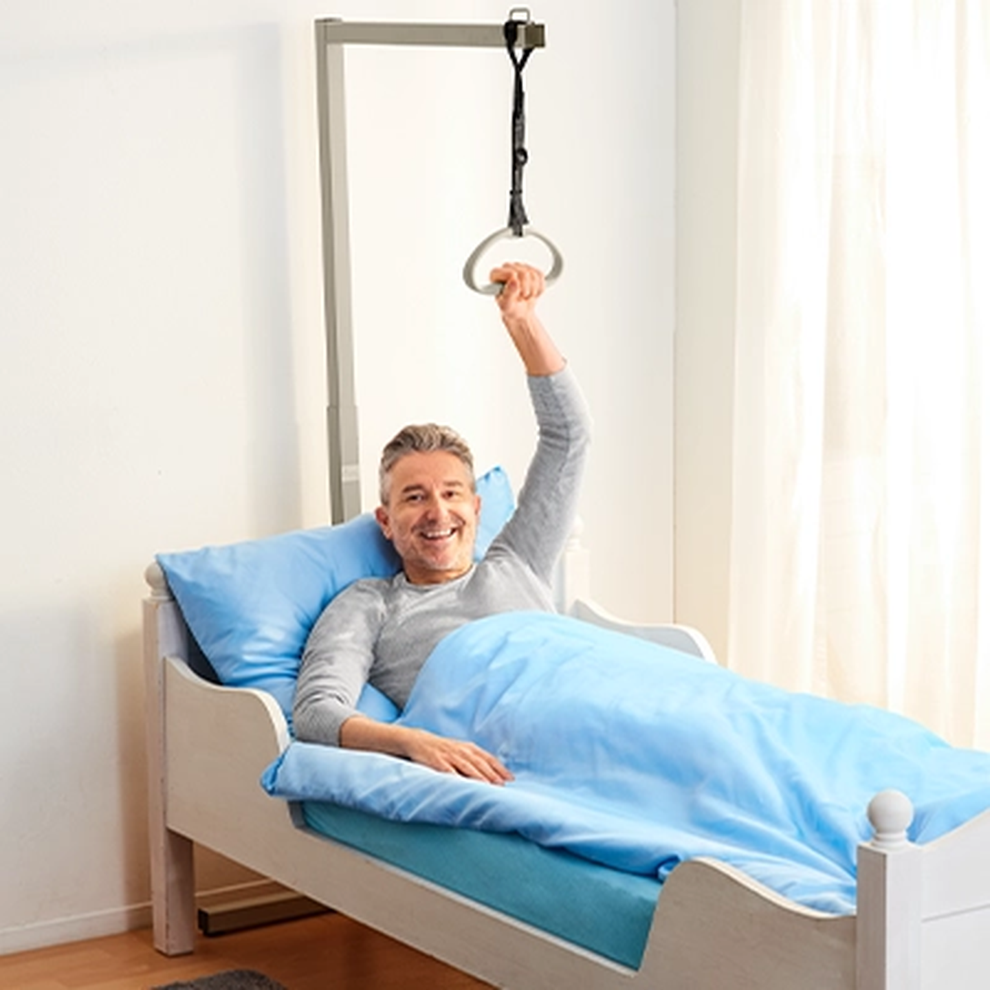 Ein lächelnder älterer Mann im Bett hält sich an einer Aufrichthilfe, in einem Zimmer mit hellblauer Bettwäsche und hellen Vorhängen.