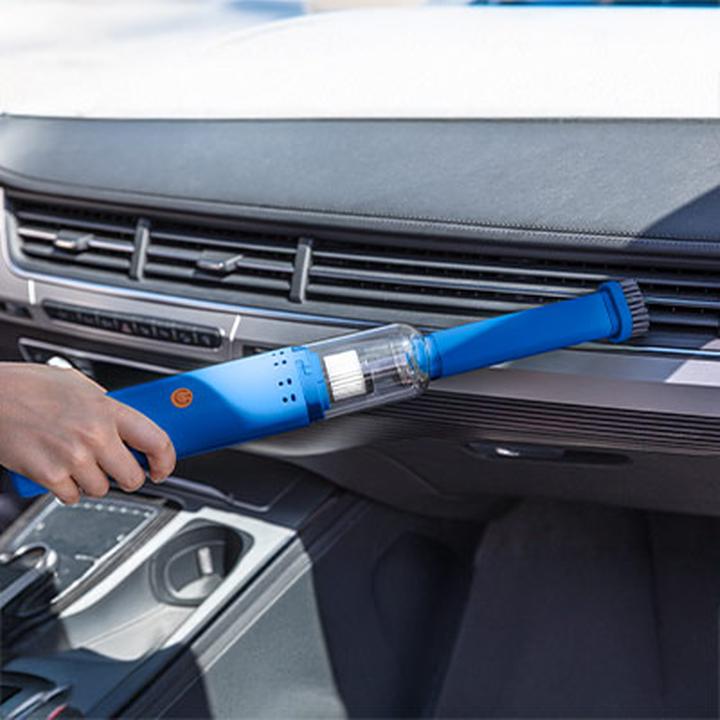 Eine Hand, die mit einem blauen Handstaubsauger Krümel von einer Holzplatte entfernt. Eine Person, die mit einem blauen Handstaubsauger die Lüftungsschlitze des Armaturenbretts eines Autos reinigt und sich dabei auf die Staubentfernung konzentriert.