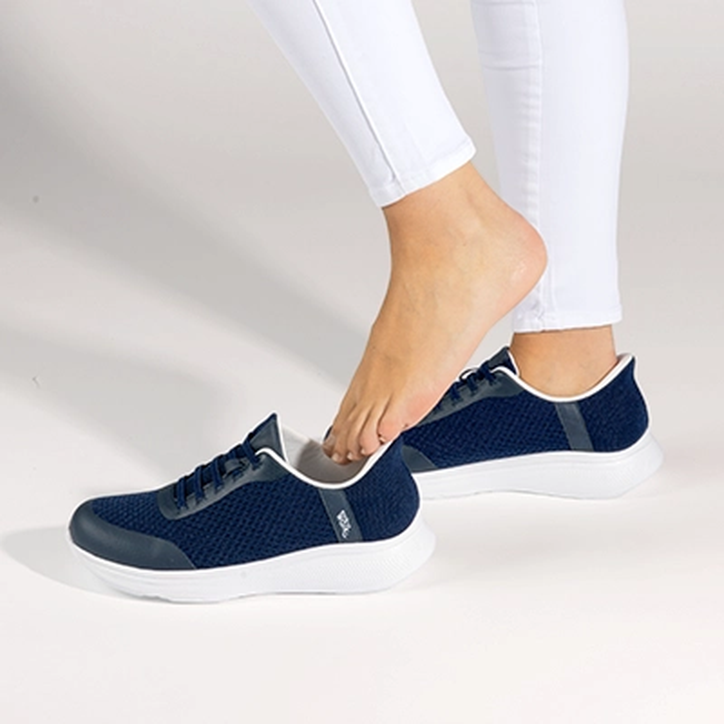 Eine Person in weißen Leggings schlüpft vor einem hellen Hintergrund in ein Paar marineblaue Sneakers mit weißen Sohlen.