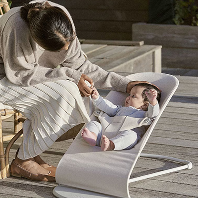 Eine Frau interagiert sanft mit einem Baby, das draußen auf einer Holzterrasse in einer beigen Babywippe liegt.