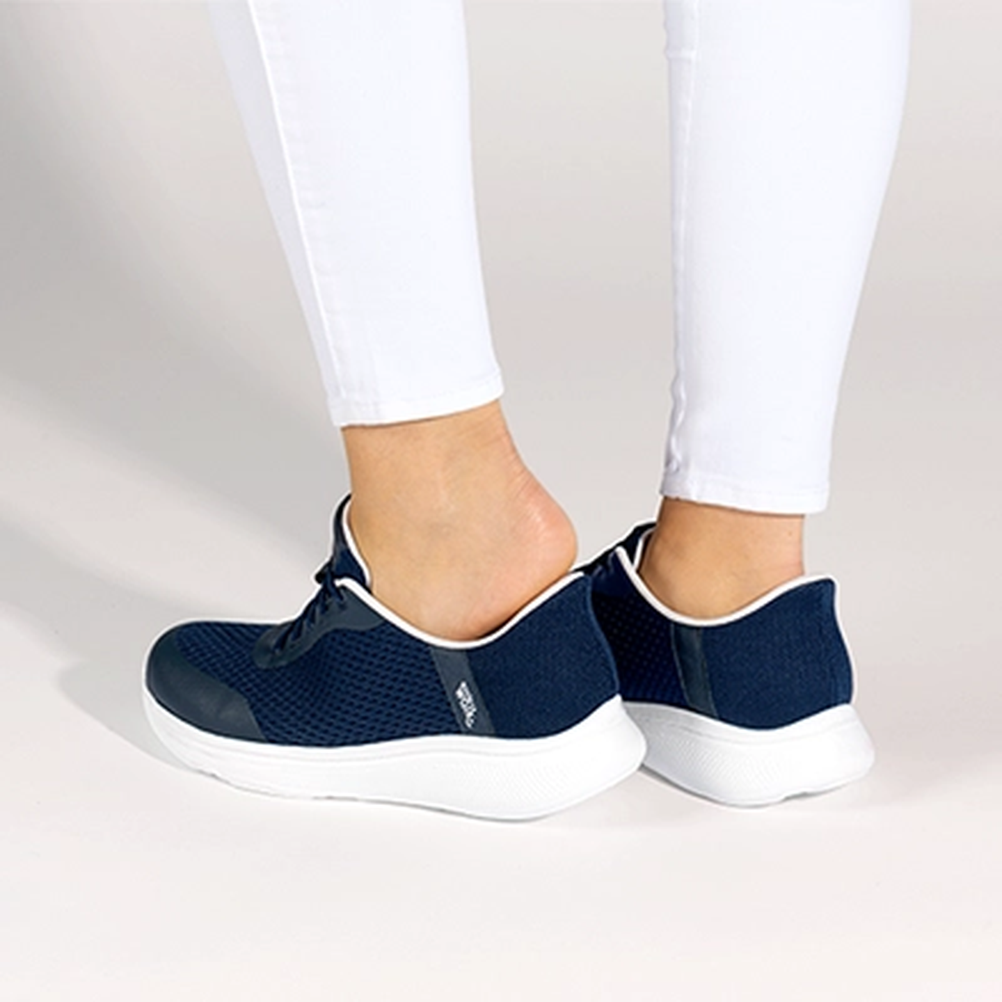 Eine Person in weißen Leggings schlüpft vor einem hellen Hintergrund in ein Paar marineblaue Sneakers mit weißen Sohlen.