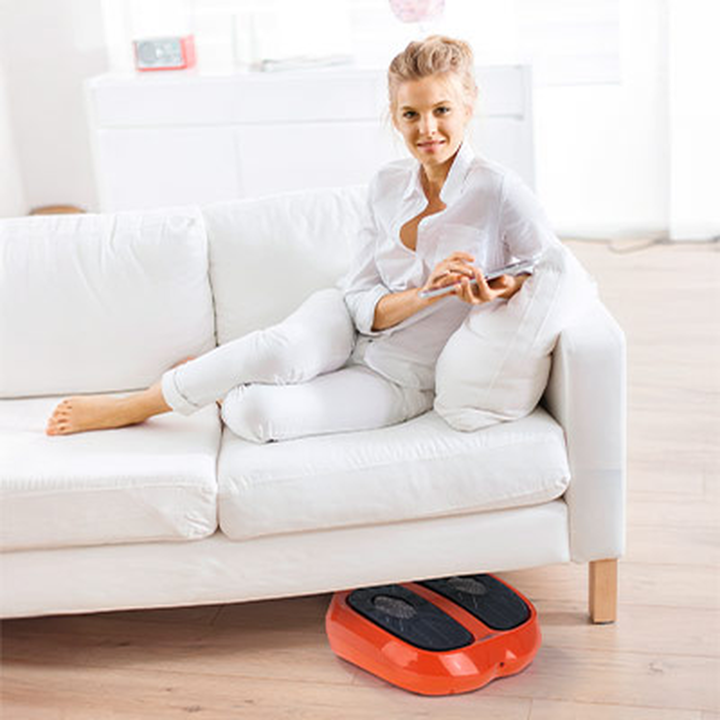 Frau in Weiß, die sich auf einem Sofa entspannt und ein rotes Fußmassagegerät auf einem Holzboden in einem hellen Raum benutzt.