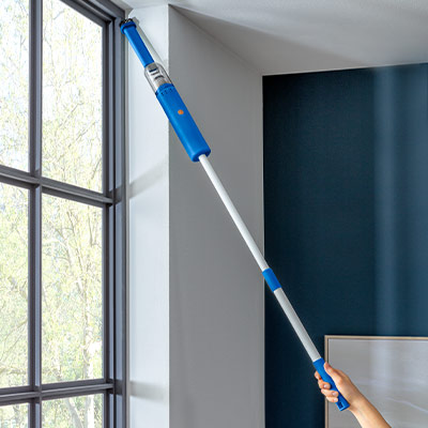 Person, die einen ausziehbaren Staubwedel mit blauem Griff benutzt, um eine hohe Ecke in der Nähe eines großen Fensters zu reinigen.