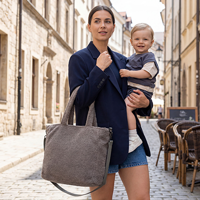 Eine Frau in einem blauen Blazer hält ein lächelndes Kind auf einer Kopfsteinpflasterstraße und trägt eine strukturierte graue Tasche.