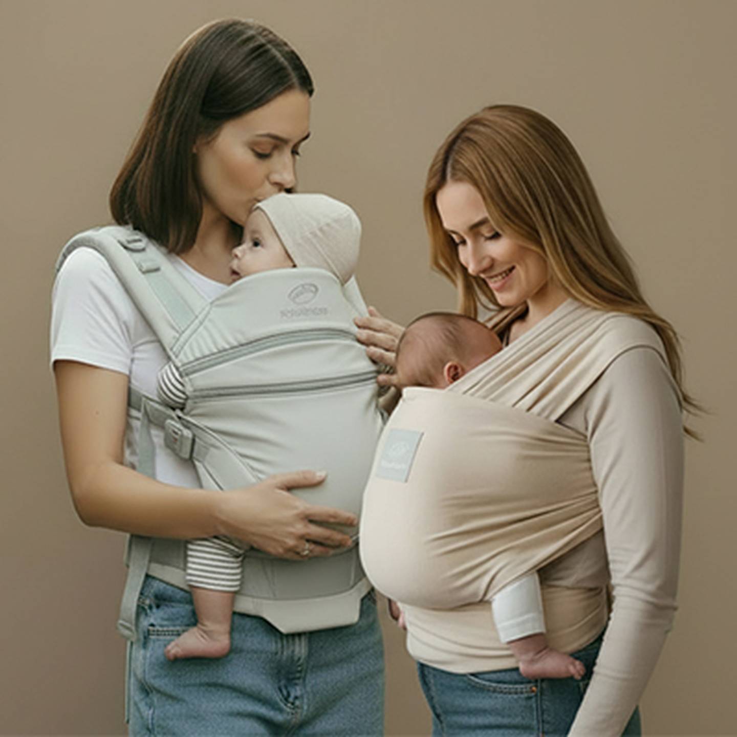 Zwei Frauen halten Babys in Tragehilfen – eine in einer grauen Trage und die andere in einem beigen Tragetuch – vor einem schlichten Hintergrund.