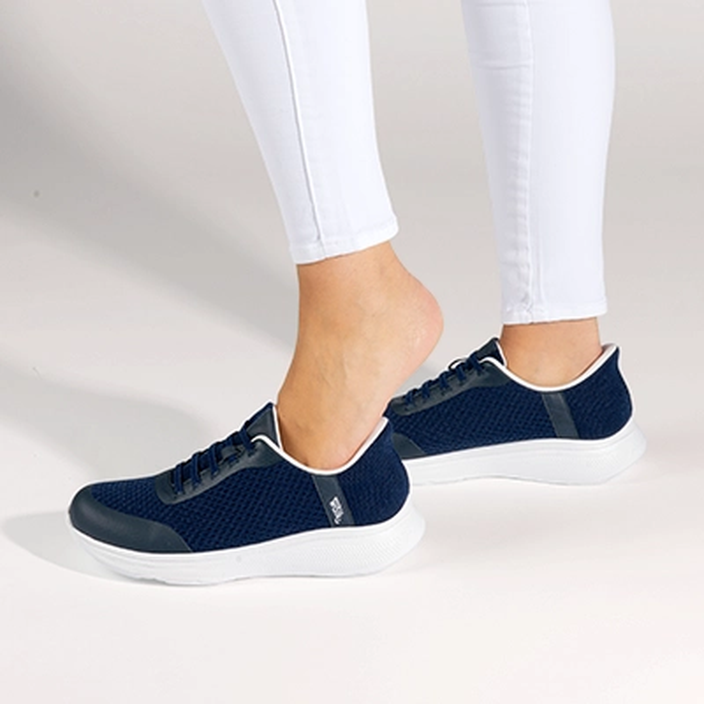 Eine Person in weißen Leggings schlüpft vor einem hellen Hintergrund in ein Paar marineblaue Sneakers mit weißen Sohlen.