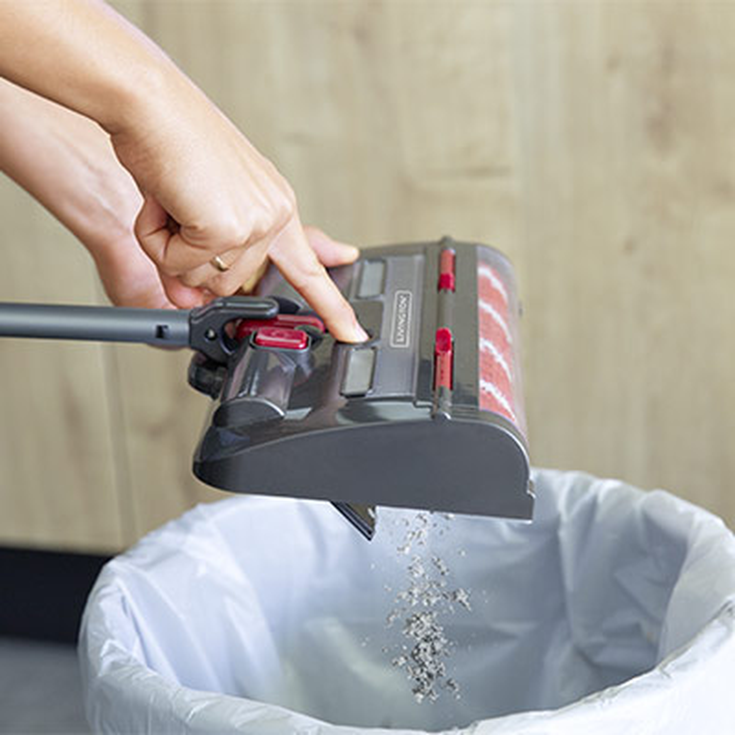 Eine Hand schüttet Schmutz aus einem kabellosen Staubsauger in einen Mülleimer. Der Staubsauger verfügt über einen roten Verschluss und ein zylindrisches Design vor einem hölzernen Hintergrund.